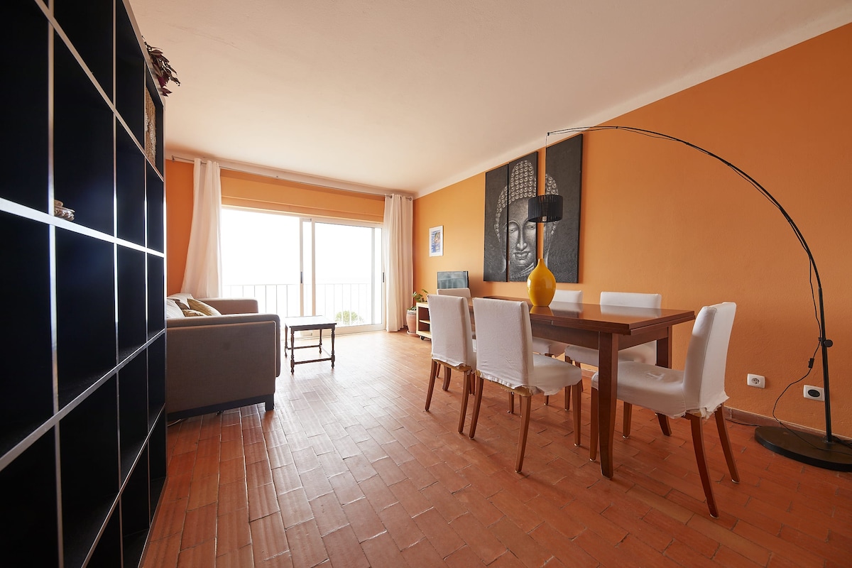 A spacious living area features warm orange walls and is furnished with a wooden dining table surrounded by white chairs. A large window offers natural light and a view, while a modern lamp and a comfortable sofa add to the inviting atmosphere.