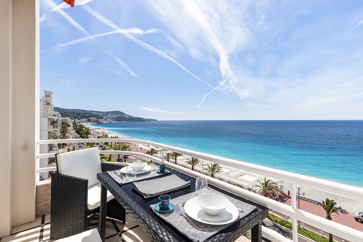 A terrace is presented with a dining table set for two, featuring neatly arranged plates and bowls. The view showcases a sweeping panorama of the coastline, with the vibrant blue sea extending to the horizon and palm trees lining the beachfront below.