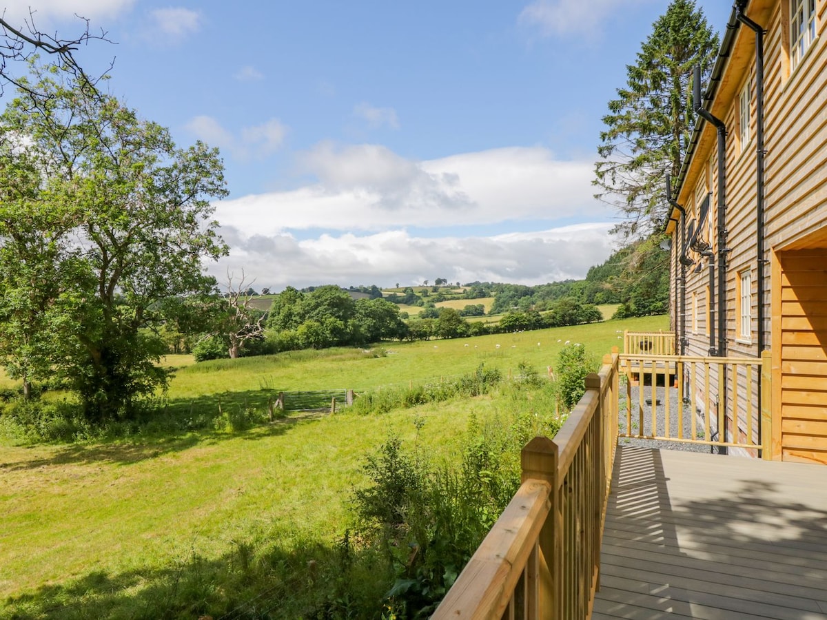 A private decking area extends from the cottage, providing an unobstructed view of the lush, rolling landscape. The scene features grazing animals in the distance, surrounded by green fields and scattered trees under a partly cloudy blue sky.