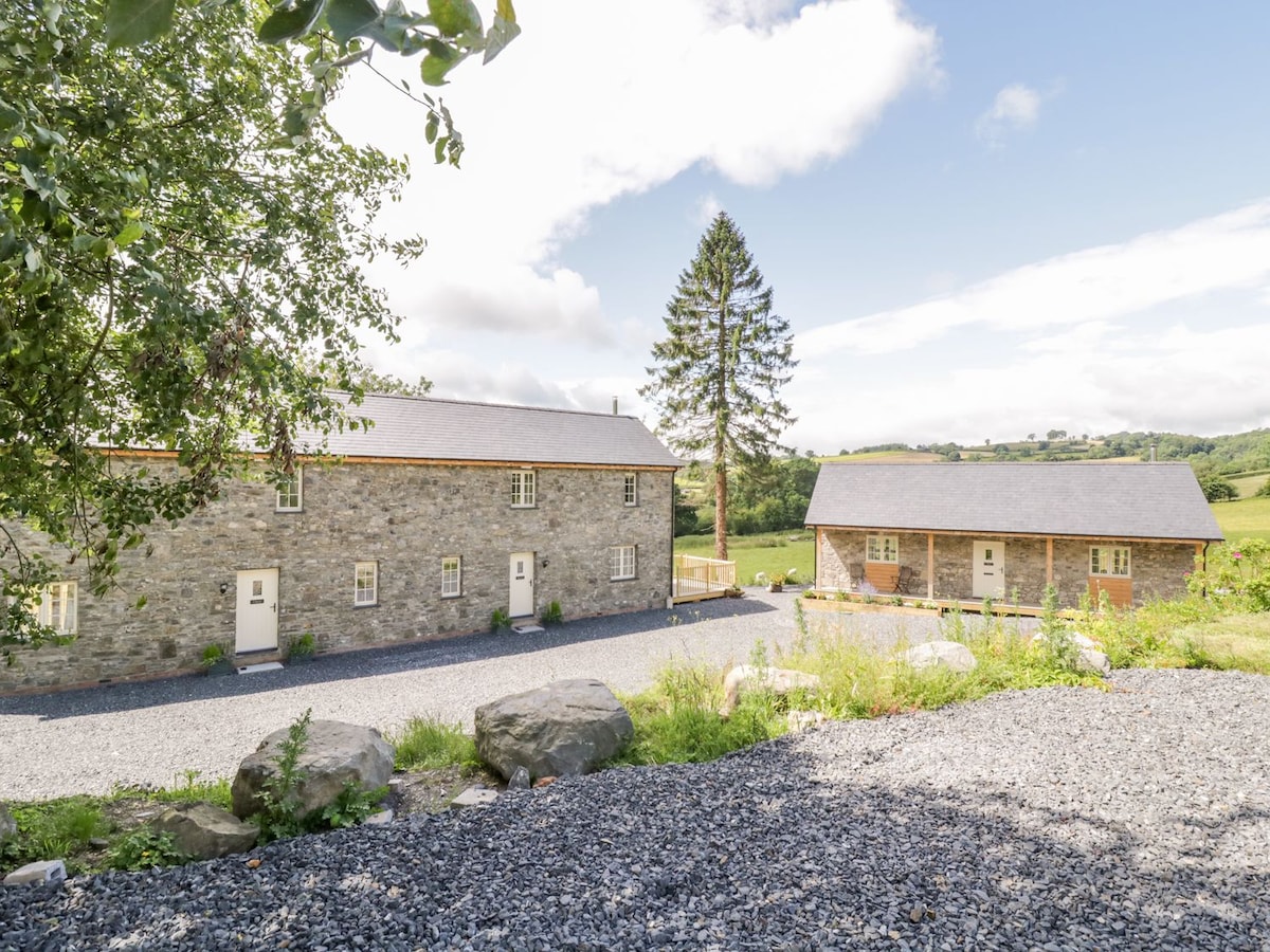 Two charming stone cottages are visible, surrounded by lush greenery. The cottages feature multiple doorways and windows, with a gravel pathway leading up to them. A tall tree stands prominently in the background, adding to the serene countryside setting.