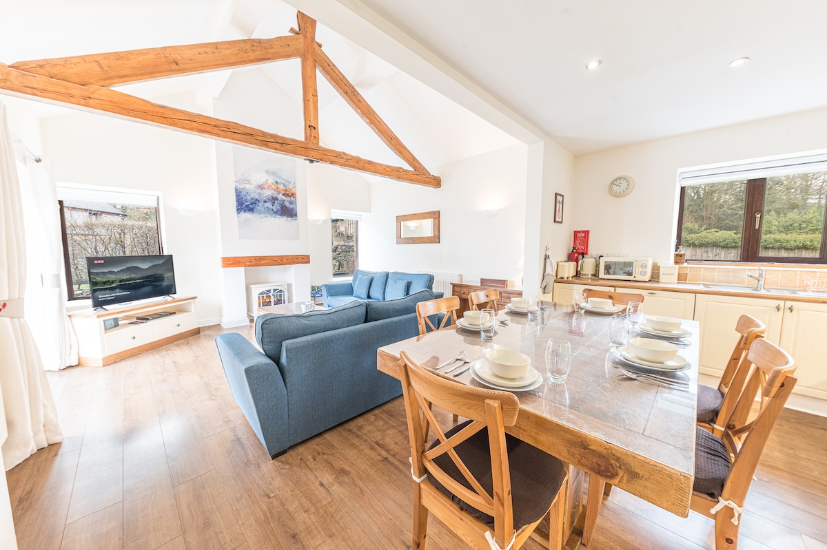 An open plan living and dining area features exposed wooden beams and large windows letting in natural light. A cozy seating arrangement in a soothing blue hue is complemented by a wooden dining table set for guests. New World kitchen appliances are visible in the background.