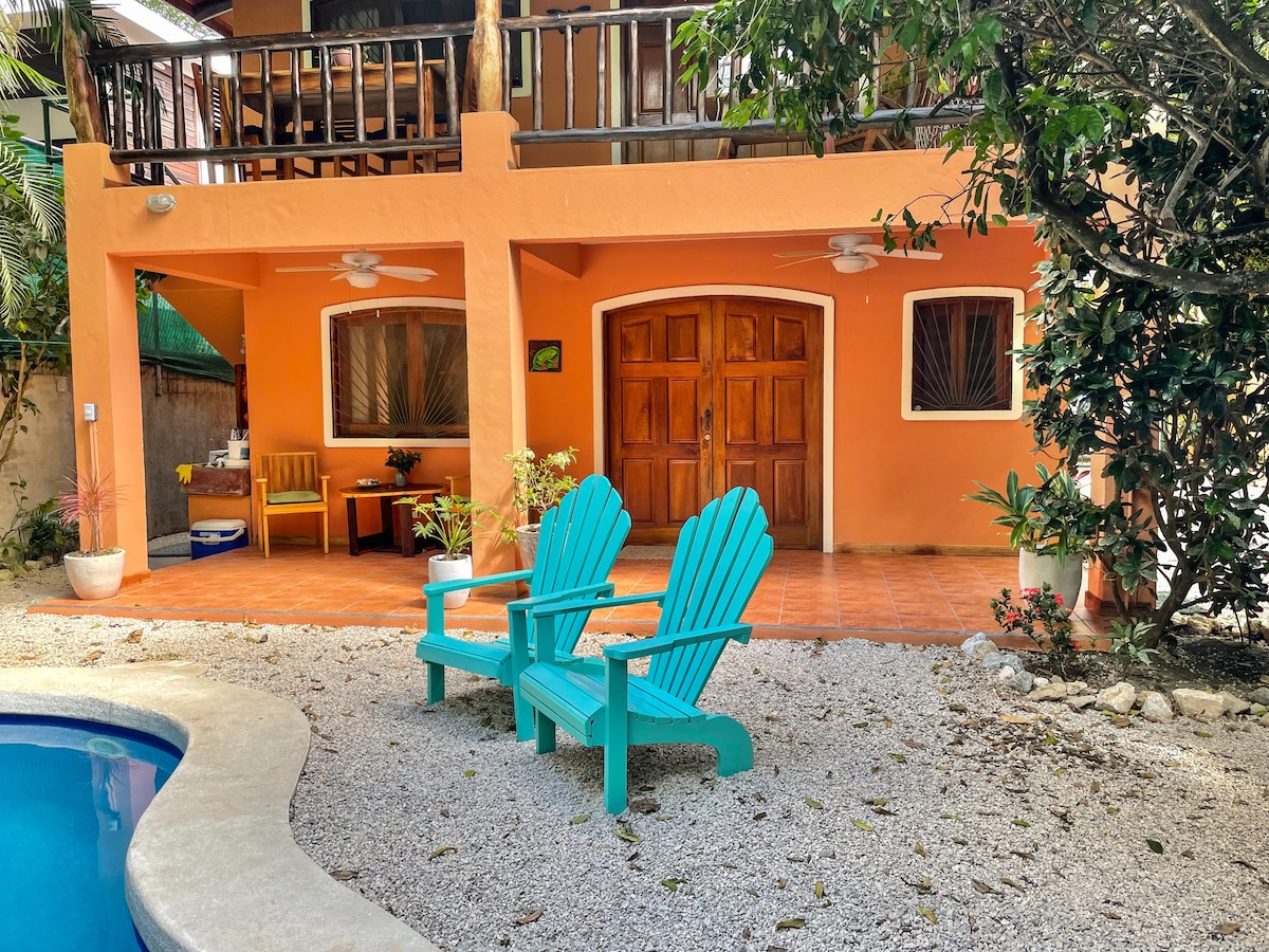 Brightly colored walls frame the entrance to the house, featuring two wooden doors and windows with shutters. Two turquoise adirondack chairs are positioned near a pool, surrounded by pebbled landscaping and tropical plants, creating a relaxed outdoor space.