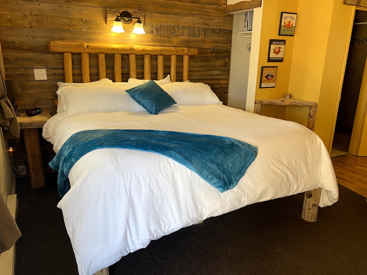 A spacious bed is framed by custom woodwork and illuminated by warm lighting. The bed features crisp white linens, complemented by a teal throw pillow and a matching blanket. Rustic wooden accents create a cozy and inviting atmosphere in the room.