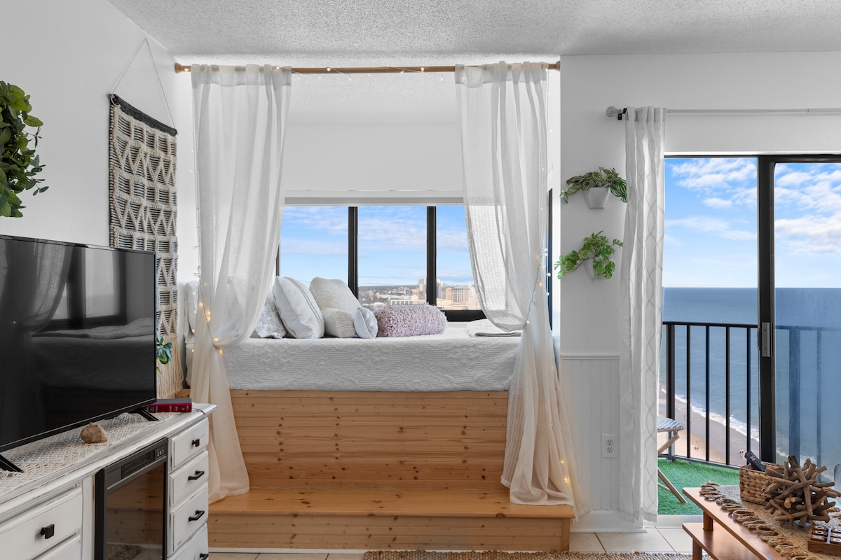 A cozy nook features a queen size bed with soft bedding, illuminated by natural light from large windows. Sheer curtains drape elegantly, enhancing the ocean view. A TV and decorative elements add a touch of charm to the inviting space.