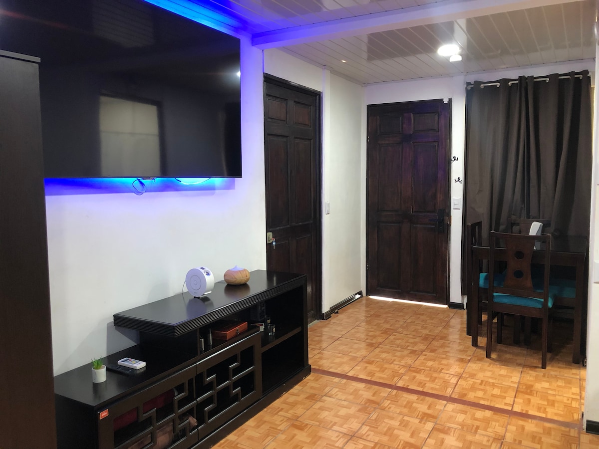 A living area is shown featuring a large flat-screen television mounted on a wall. A dark wooden dining table with matching chairs is visible, complemented by blue accent lighting under a shelf. The space is well-lit by ceiling fixtures, and two wooden doors lead to other areas.