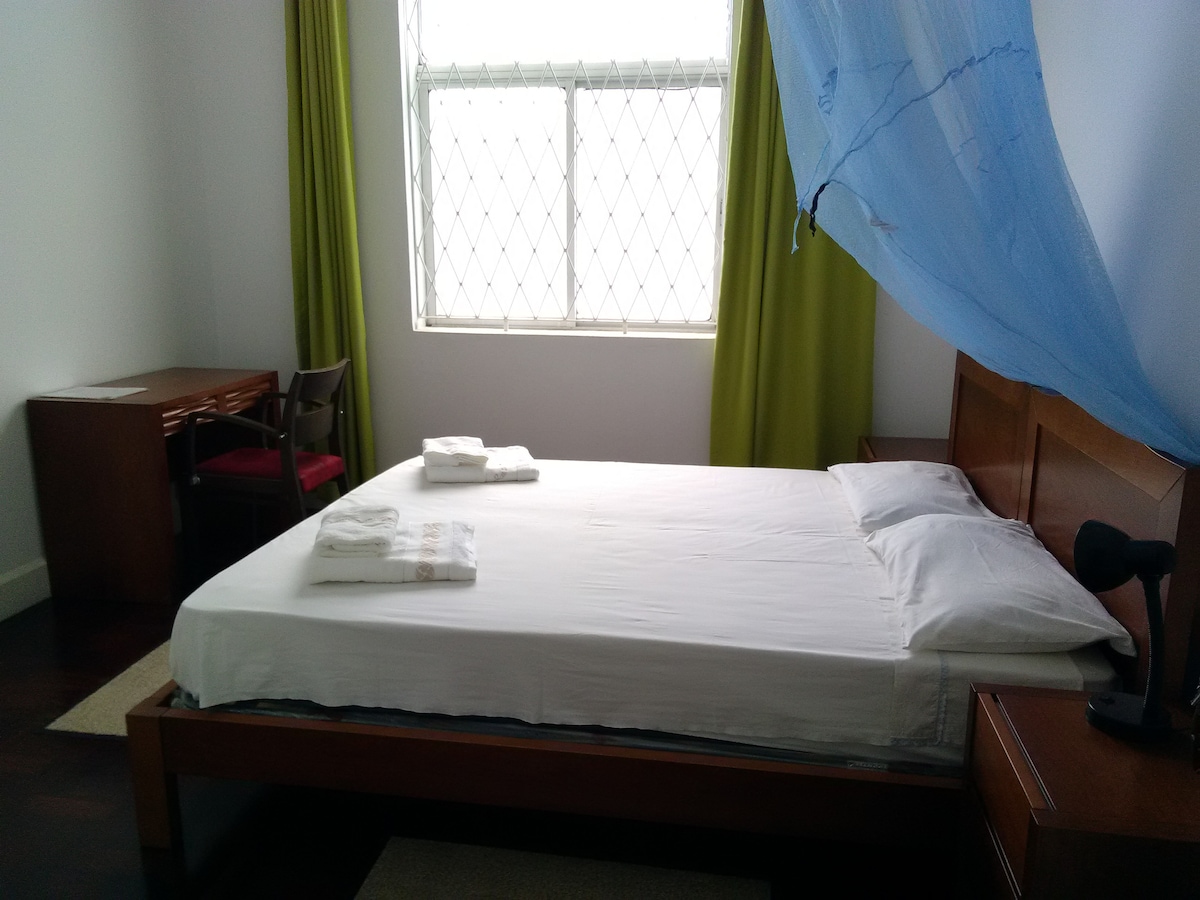 A bright and airy bedroom features a spacious bed with freshly laundered white linens. A work desk with a chair is positioned against the wall. Natural light filters through a large window with diamond-patterned panes, and green curtains add a touch of color.