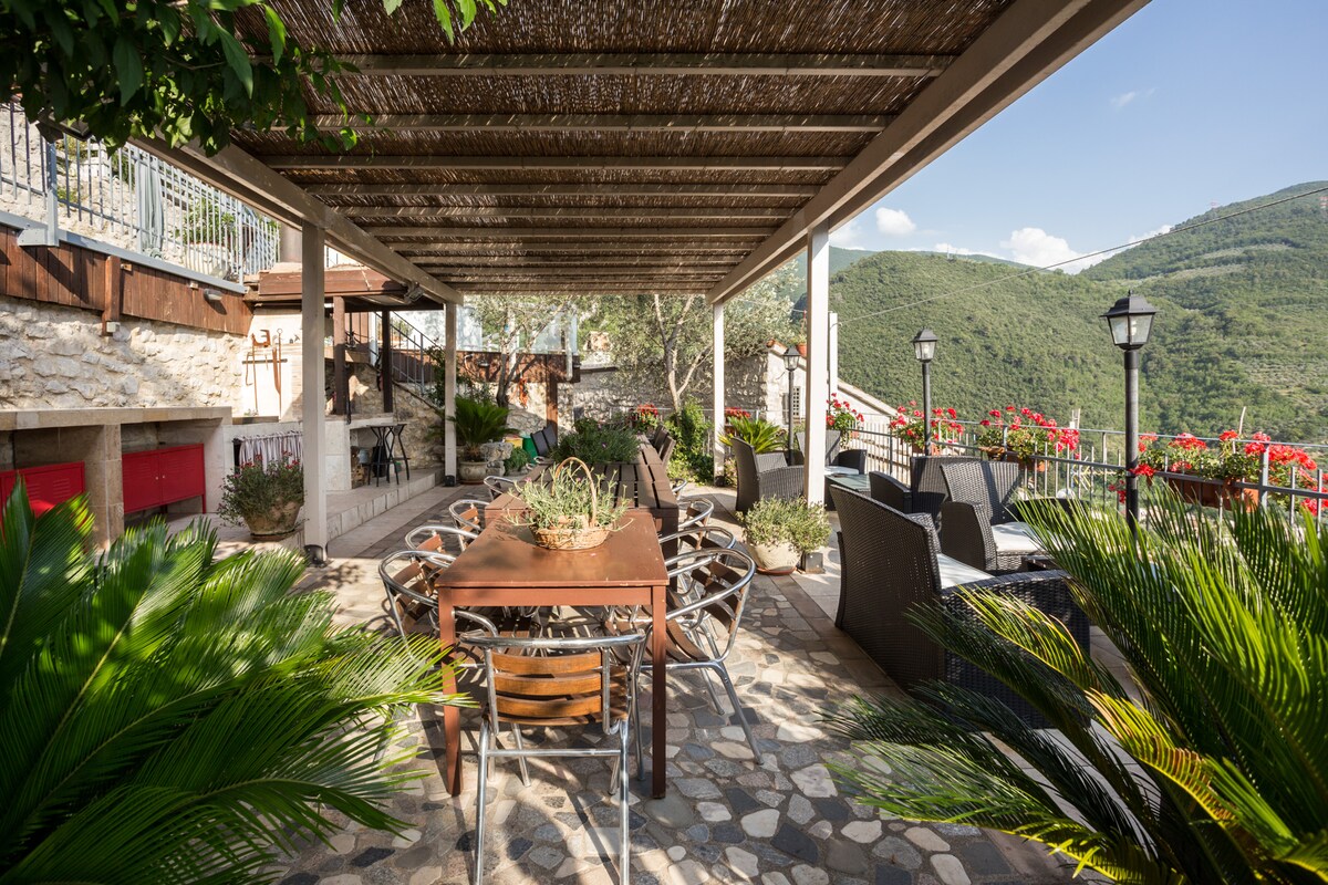 An outdoor dining area is framed by a wooden pergola, providing shade. A large wooden table with surrounding chairs invites gatherings. Lush greenery and flowering plants enhance the setting, while scenic mountains are visible in the background, creating a peaceful atmosphere.