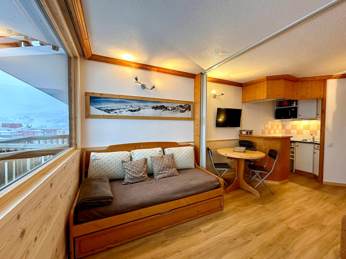 A bright living area features a cozy daybed adorned with decorative pillows. A round dining table is surrounded by four chairs. A flat-screen TV is mounted on the wall, and the kitchen area is visible in the background, showcasing wooden cabinetry.