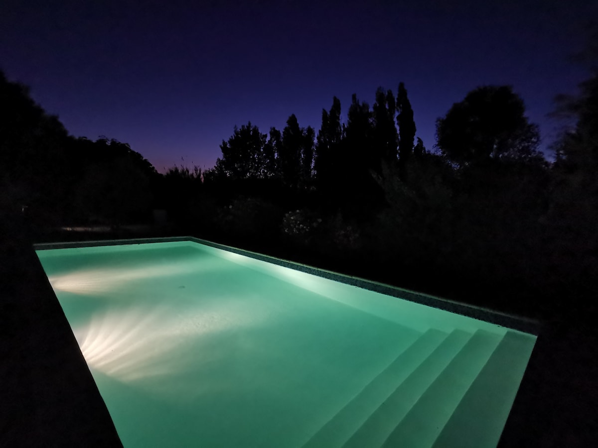 A peaceful swimming pool is illuminated in soft turquoise hues under the night sky. Gentle steps lead into the water, surrounded by lush greenery, creating a serene outdoor space.