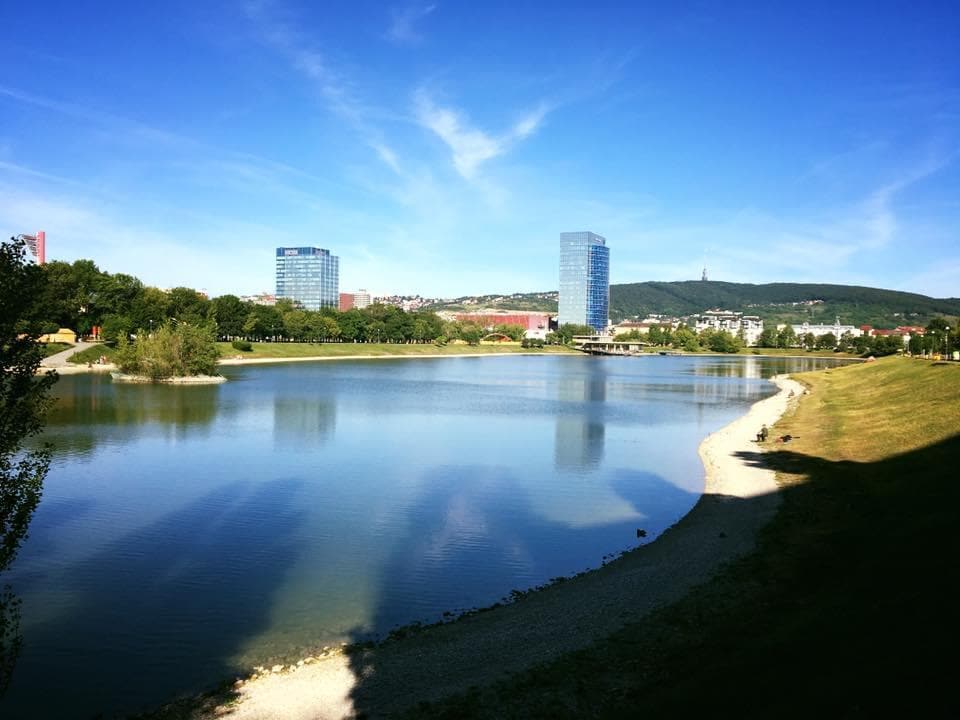 A calm lake reflects the blue sky, bordered by a green shoreline. Modern buildings are visible in the background, while trees provide shade along the water's edge. The peaceful scene invites relaxation and outdoor activities.