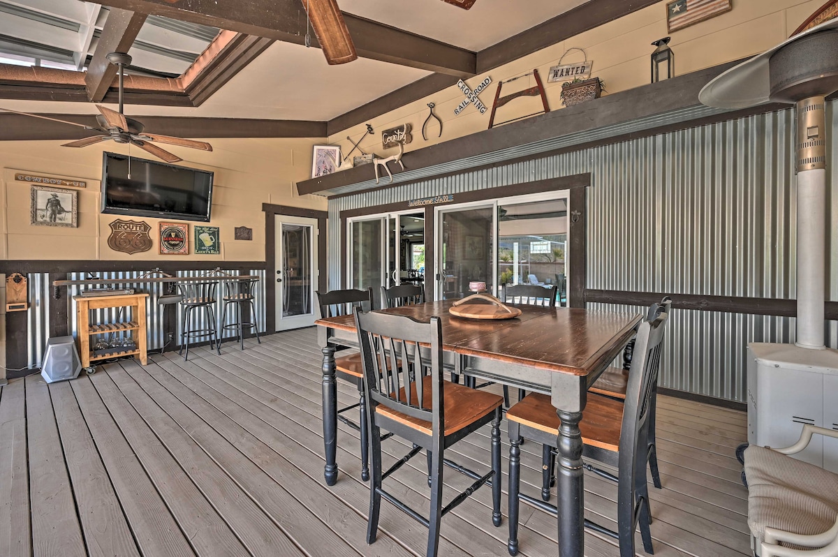 An outdoor dining area features a wooden table surrounded by black chairs. Decor includes rustic wall art and a flat-screen TV mounted above. Ceiling fans provide airflow, while a heater is positioned in the corner. Sliding glass doors lead to the interior.