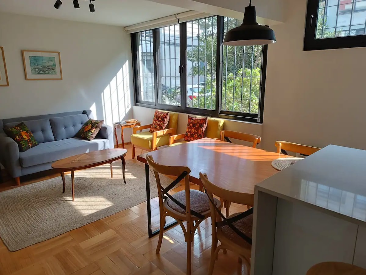 A bright and airy living area is highlighted, featuring a comfortable gray sofa and two yellow chairs with colorful cushions. A wooden coffee table sits center stage, surrounded by a dining table and chairs. Natural light filters through large windows, illuminating the inviting space.