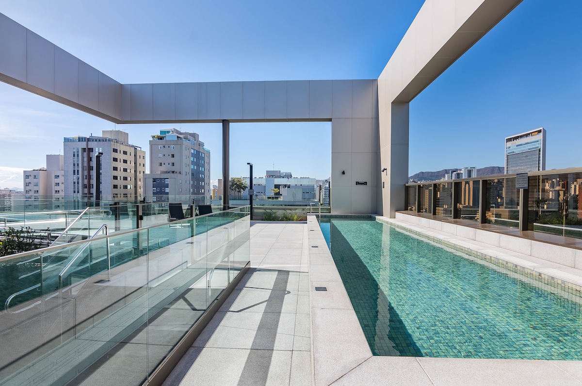 A modern rooftop terrace features a rectangular swimming pool surrounded by clean lines and glass railings. Sun loungers are positioned along the pool edge, with a skyline view of the city in the background. The area is illuminated by natural light.