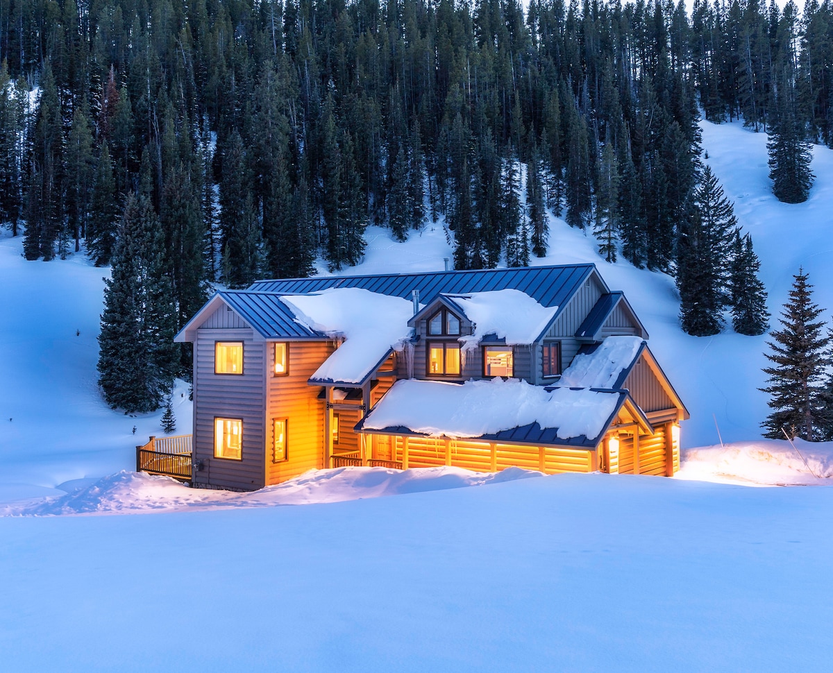 The exterior of Long Lake Lodge is showcased with a covered porch and multiple windows glowing warmly against a snowy backdrop. The two-story structure is surrounded by evergreen trees, with substantial snow accumulation on the roof and ground, creating a serene winter landscape.