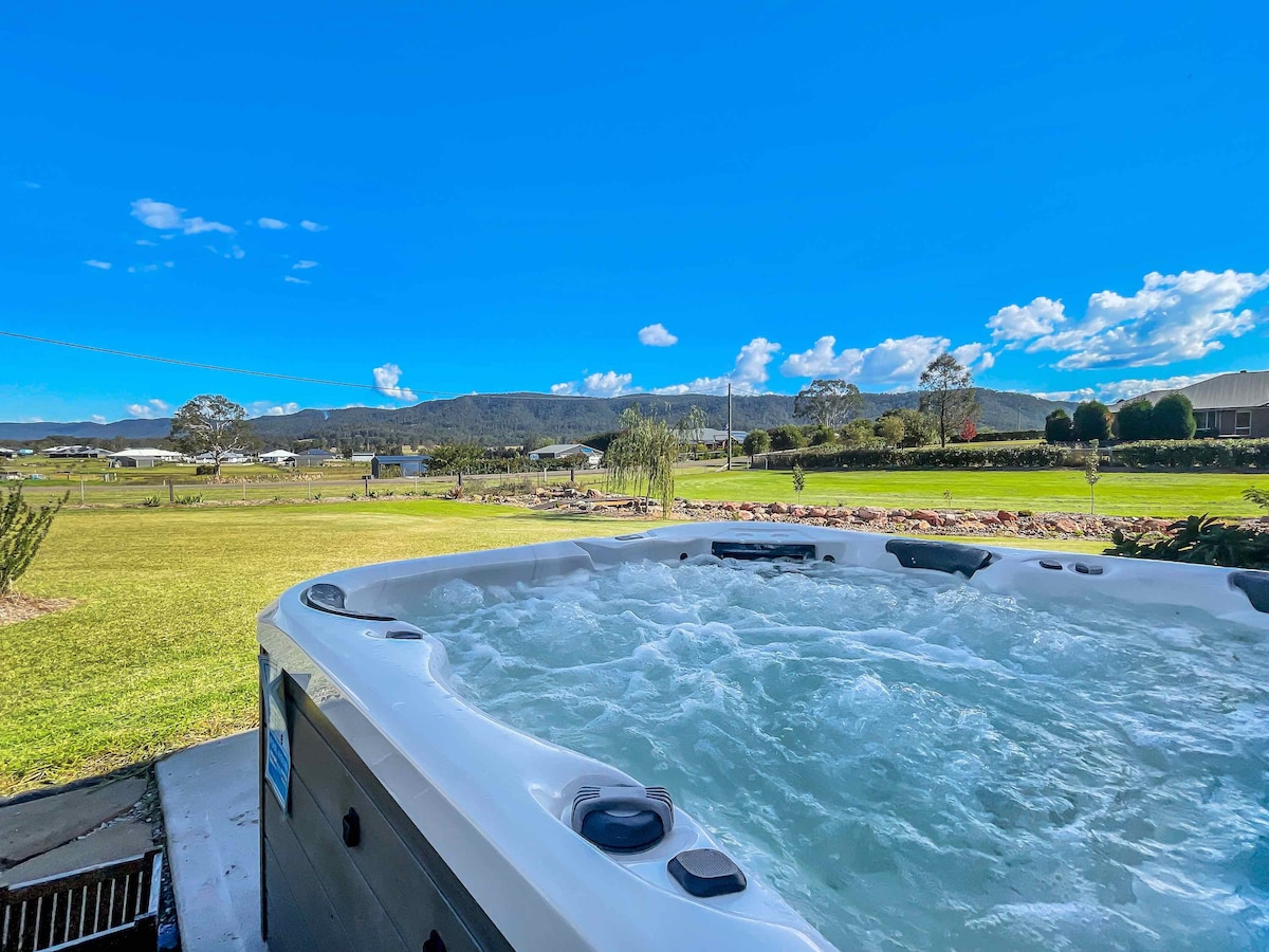 A spacious outdoor spa bubbles invitingly under a clear blue sky. The surrounding landscape features lush green grass and distant mountains, creating a serene setting. Soft clouds drift by, enhancing the peaceful atmosphere of the area.