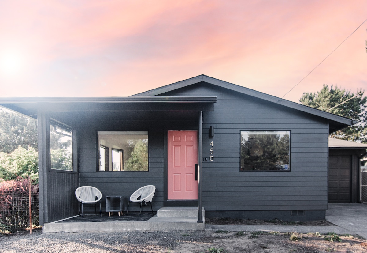 The exterior of a modern bungalow is shown, featuring a welcoming pink front door and large windows. Two stylish chairs with a small table are positioned on the porch. The structure is painted in a dark hue, surrounded by greenery and a gravel pathway.