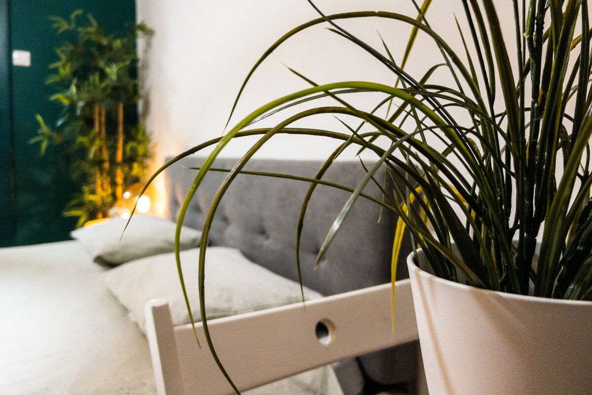 A cozy bedroom corner features a spacious bed dressed in neutral tones, complemented by soft pillows. A green plant adds a touch of nature, while warm lighting creates a calming atmosphere in the background. The overall space is designed for relaxation.