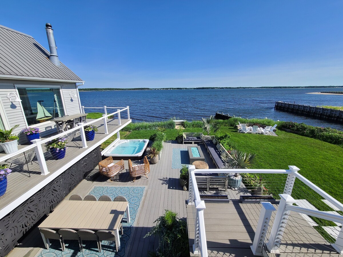 A serene waterfront view is captured from the deck, showcasing a well-appointed outdoor space that includes a hot tub, dining area, and lounge chairs. The green lawns and waterway create a tranquil setting, inviting relaxation amidst nature.