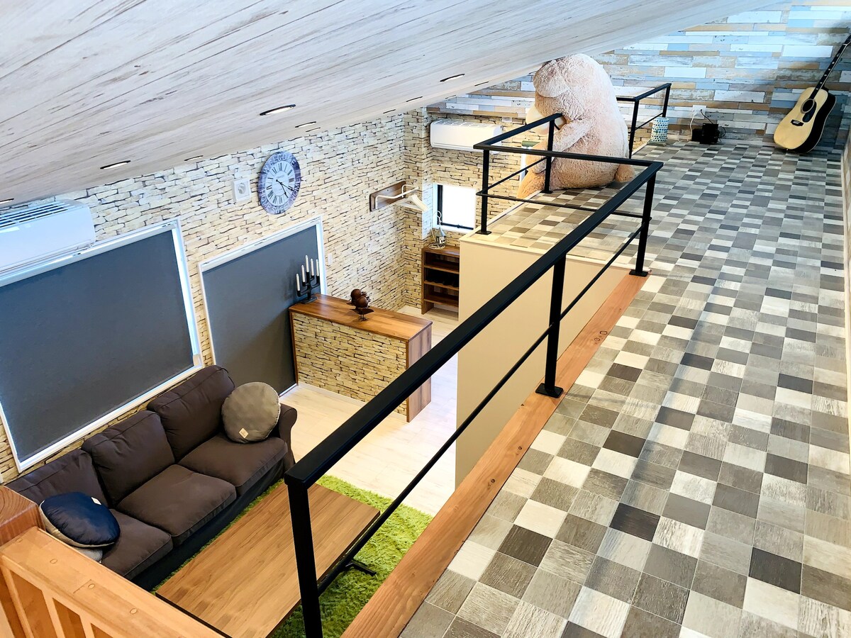 An open loft space displays a cozy seating area with a dark sofa and a large wall clock. A plush bear stands in a corner, while a checkered floor extends throughout the area. Green accents from a rug add a touch of color.