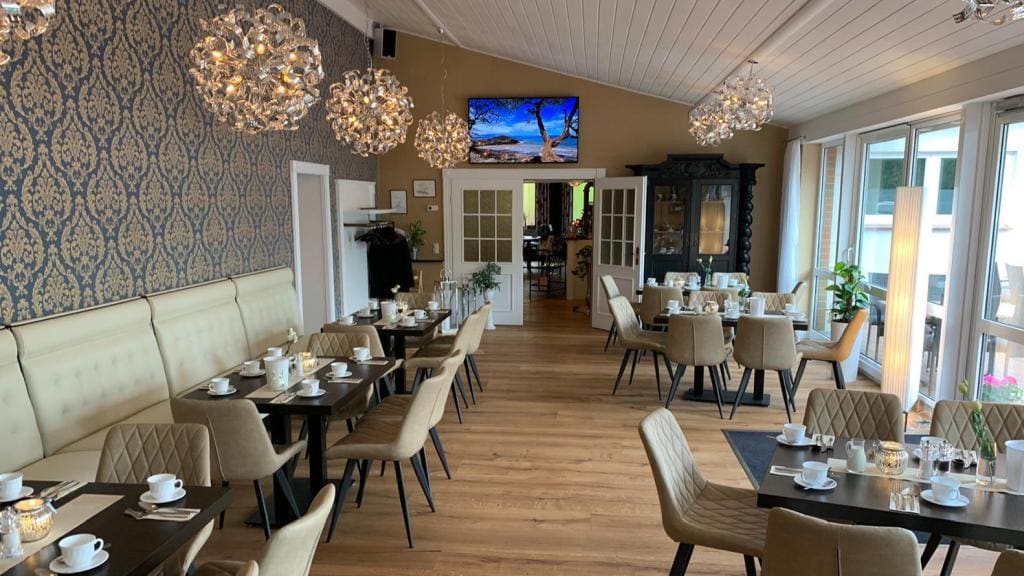 A dining area features elegant furnishings with beige upholstered seating and dark wood tables arranged for guests. Decorative light fixtures hang from the ceiling, and a large screen displays tranquil scenery. Glass doors open to a view outside, enhancing the spacious ambiance.