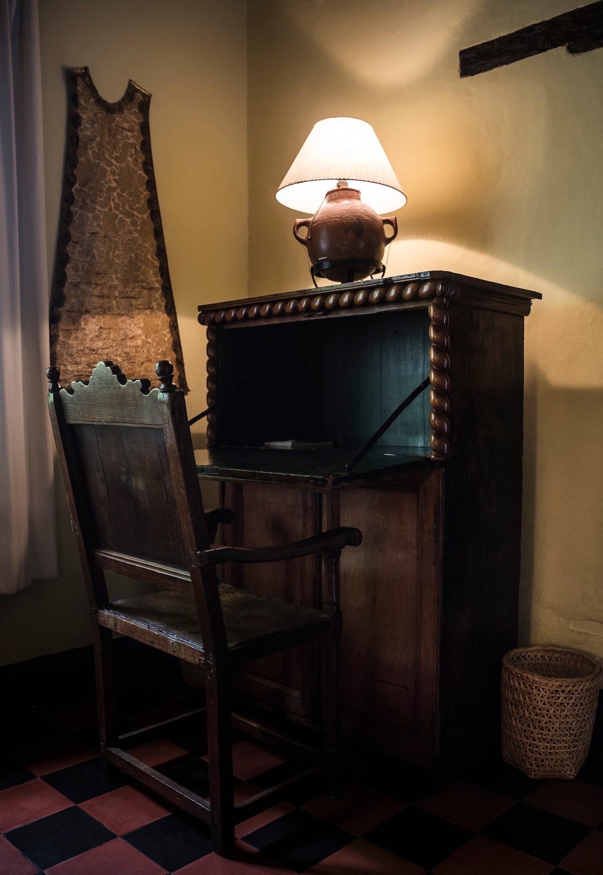 A vintage wooden desk is positioned against a wall, complemented by a high-backed chair. A soft light is provided by a lamp on the desk, creating a warm ambiance. Decorative elements include a woven basket nearby and a textured fabric backdrop.