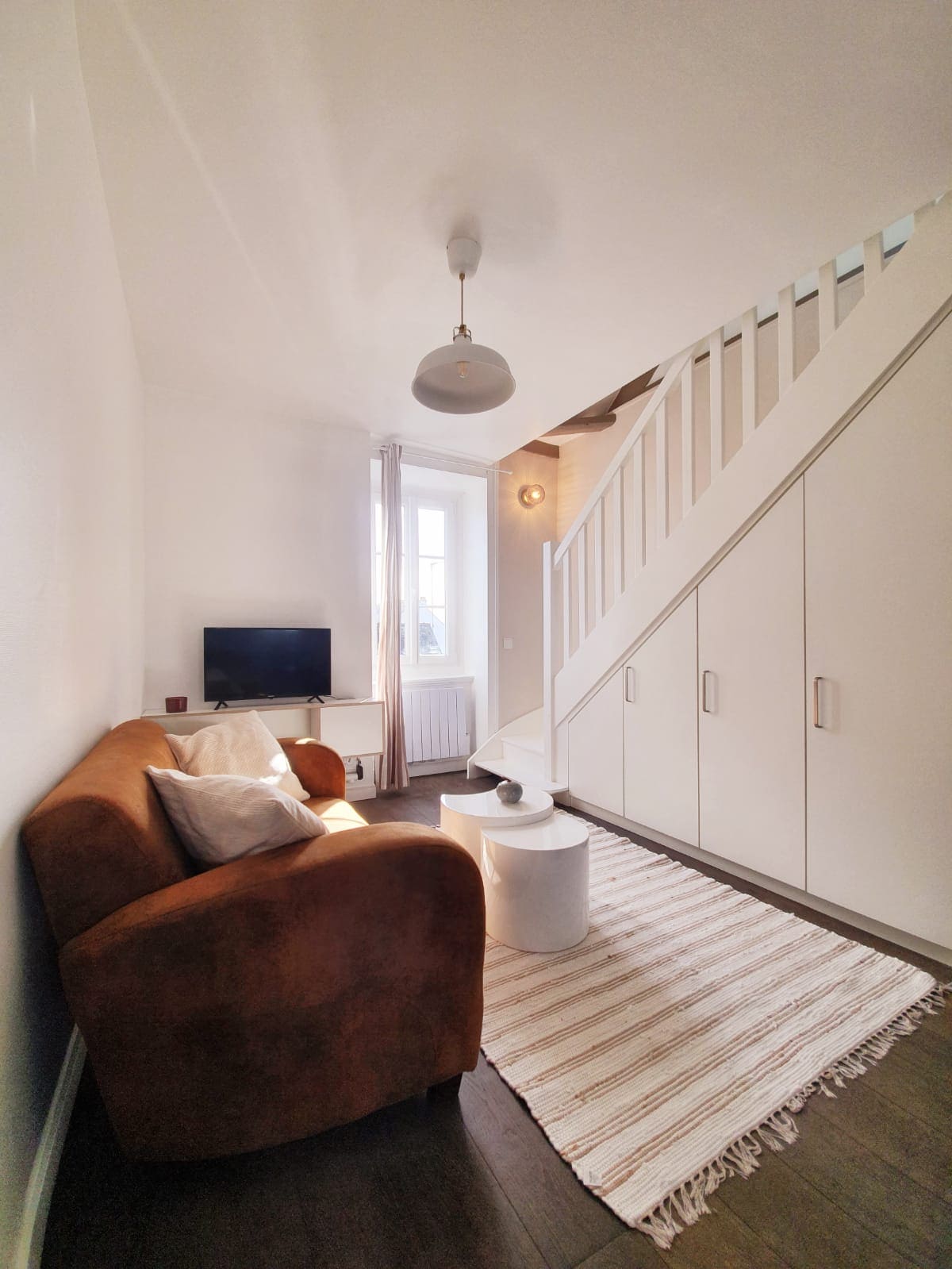 A comfortable living area is depicted with a soft sofa and a small round table. A television is mounted on the wall opposite. Natural light enters through a window beside the staircase, adding brightness to the space. White walls and light-colored furnishings create a calm ambiance.