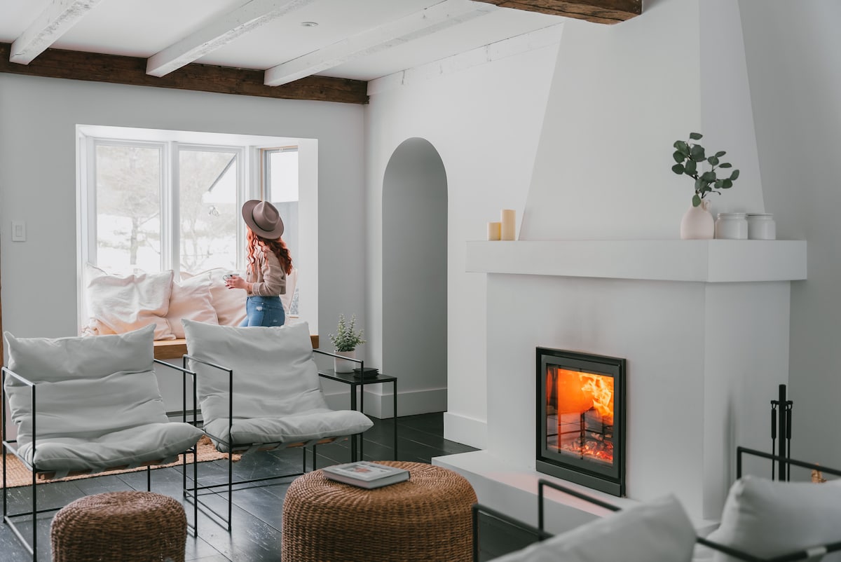 A spacious living area is highlighted by a white fireplace with a warm fire. Light-colored lounge chairs complement the room, while natural wood beams add character. A large window offers a view of the outdoor scenery, creating a bright and inviting atmosphere.