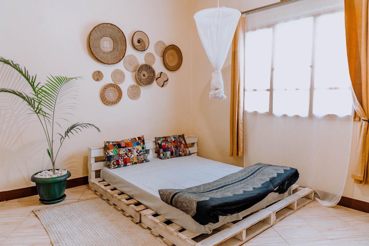 A tranquil sleeping area features a low bed made of wood pallets, adorned with colorful patterned cushions. Decorative woven wall hangings are arranged above the bed, while a potted plant adds a touch of greenery. Soft light enters through sheer curtains, creating a serene ambiance.