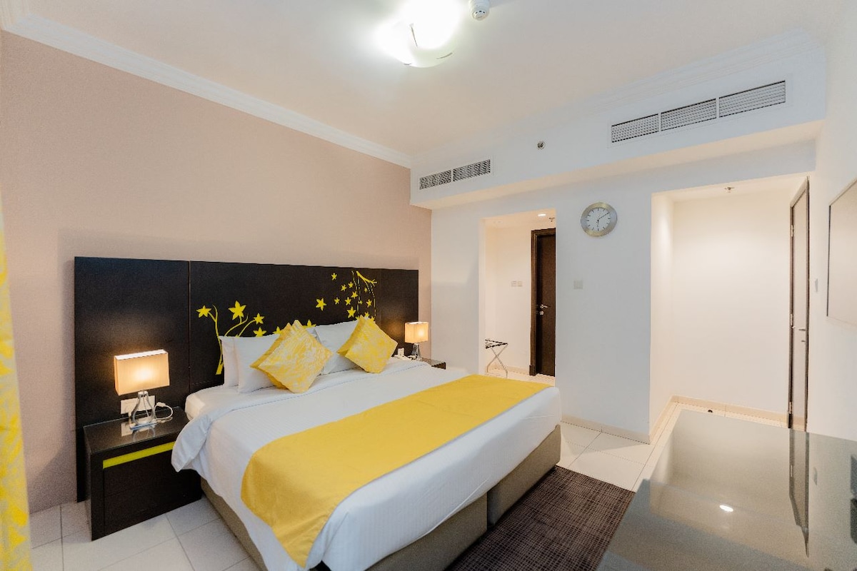 A spacious room is furnished with a large bed featuring bright yellow accents. Two bedside lamps provide soft lighting beside the bed. A modern clock is mounted on the wall, and a sleek table is positioned in the foreground, contributing to the room's contemporary design.