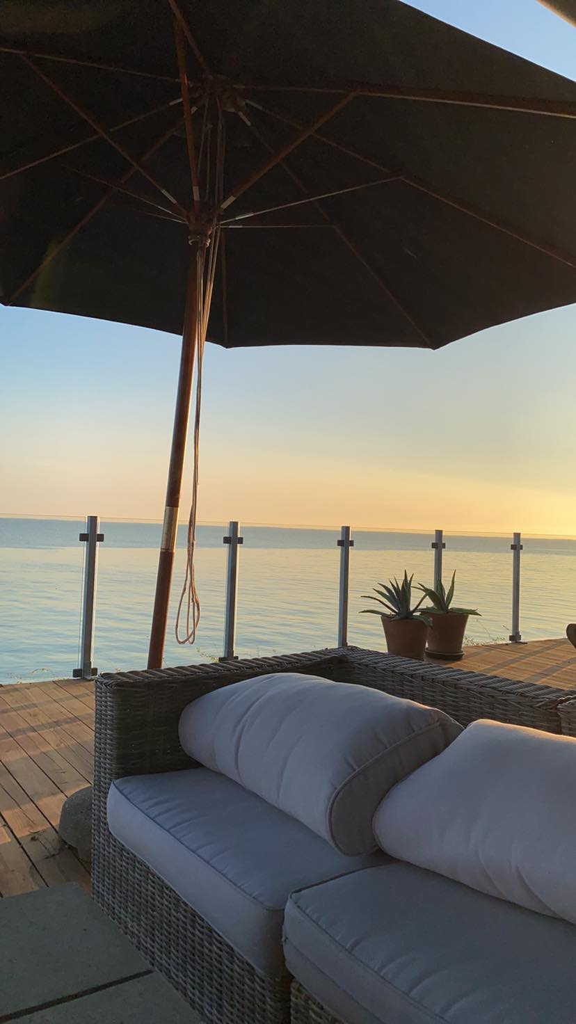 A comfortable outdoor seating area features a cozy sofa with plush cushions, positioned on a wooden deck. An umbrella provides shade while overlooking the calm sea in the background, where a serene horizon meets the sky during sunset.