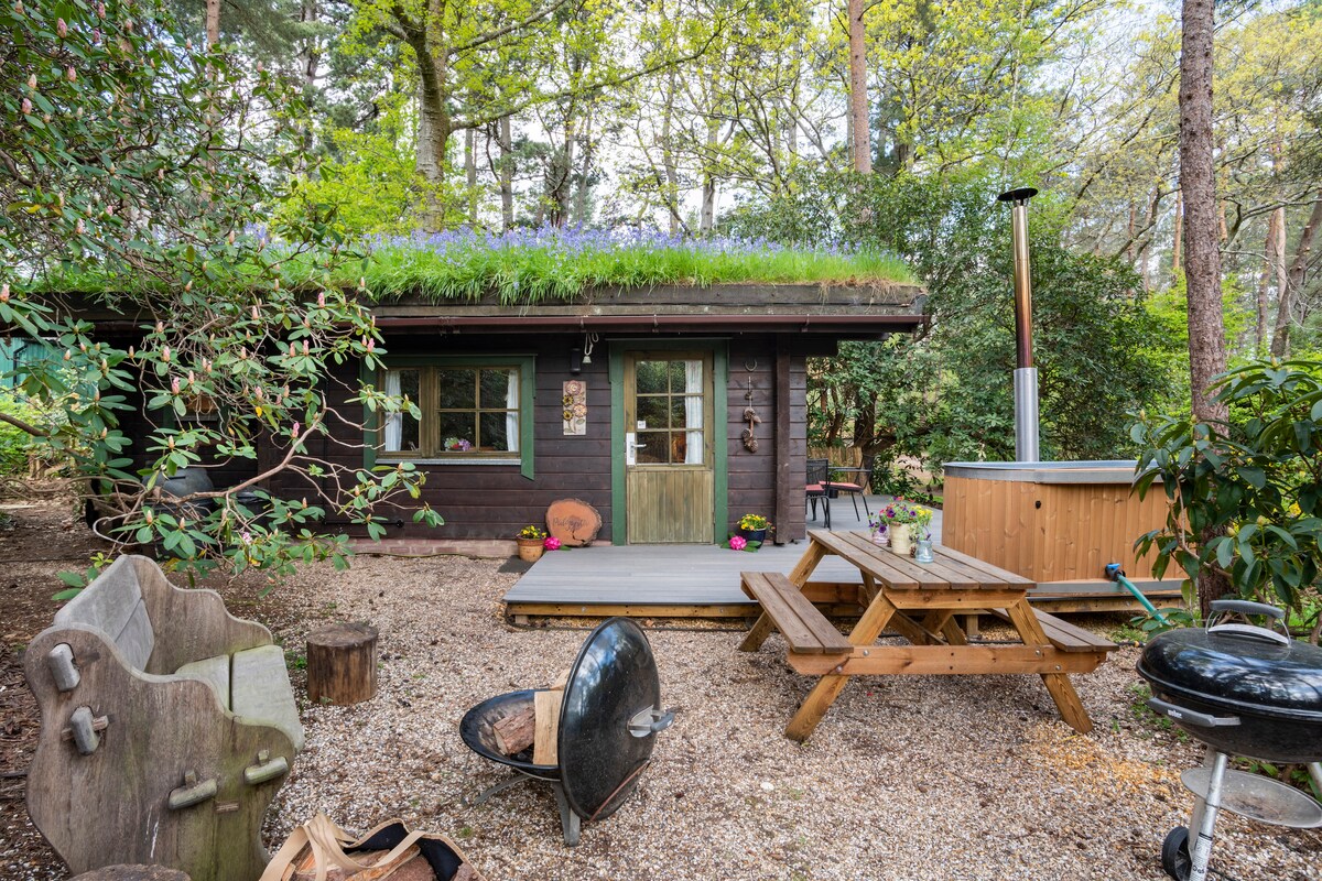 A traditional Finnish log cabin with a grass roof is surrounded by trees and rhododendrons. A wooden picnic table is positioned on a deck, complemented by outdoor seating and a barbecue. A wood-fired hot tub is visible to the side, inviting relaxation in nature.