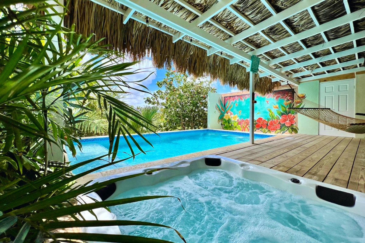 A private jacuzzi sits on a wooden deck, bubbling gently next to a sparkling blue pool. Lush greenery frames the scene, and a vibrant mural featuring tropical flowers is visible on an adjacent wall. A hammock is suspended under a thatched roof, inviting relaxation.