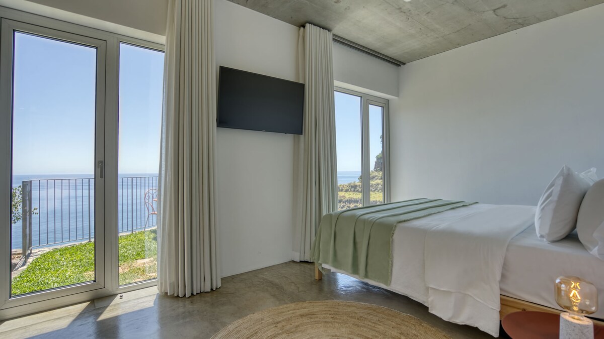 A comfortable bedroom features a spacious bed with light-colored linens. Large glass doors and windows provide views of the ocean and natural surroundings. A television is mounted on the wall, and soft curtains frame the windows, allowing natural light to enter.