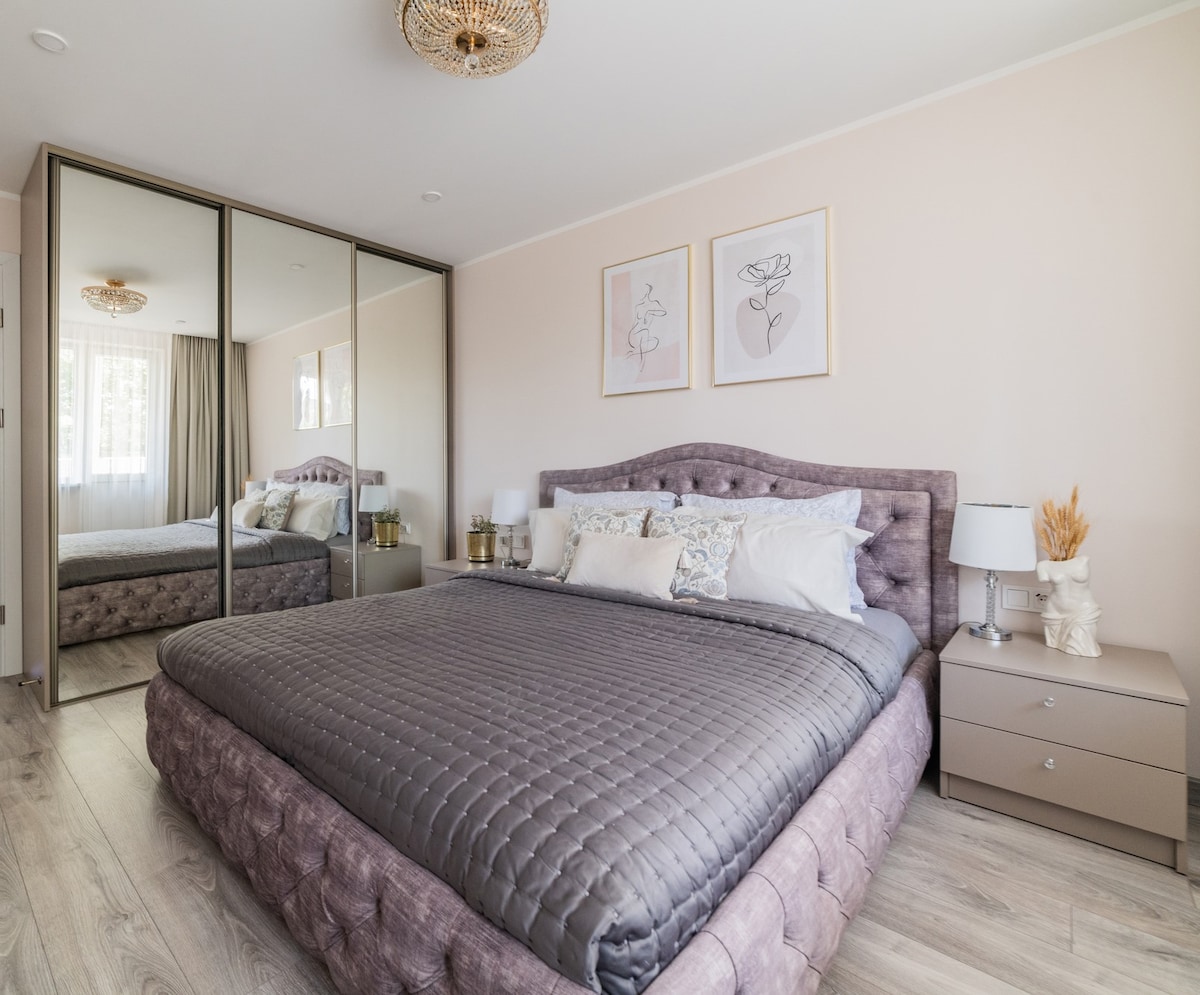A spacious bedroom features a large bed with a quilted gray coverlet. Framed artwork hangs above the headboard. Flanking the bed are modern nightstands with lamps. A mirrored wardrobe reflects the natural light coming from a nearby window, enhancing the room's brightness.