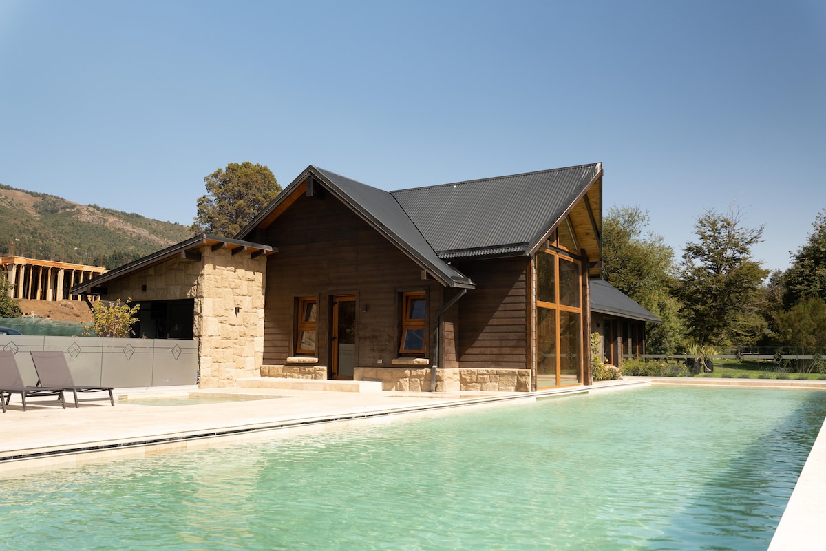 A modern house constructed with natural stone and wood features is positioned near a clear swimming pool. Large windows allow for ample light, while sun loungers are arranged beside the water's edge. A scenic view of surrounding nature enhances the outdoor space.