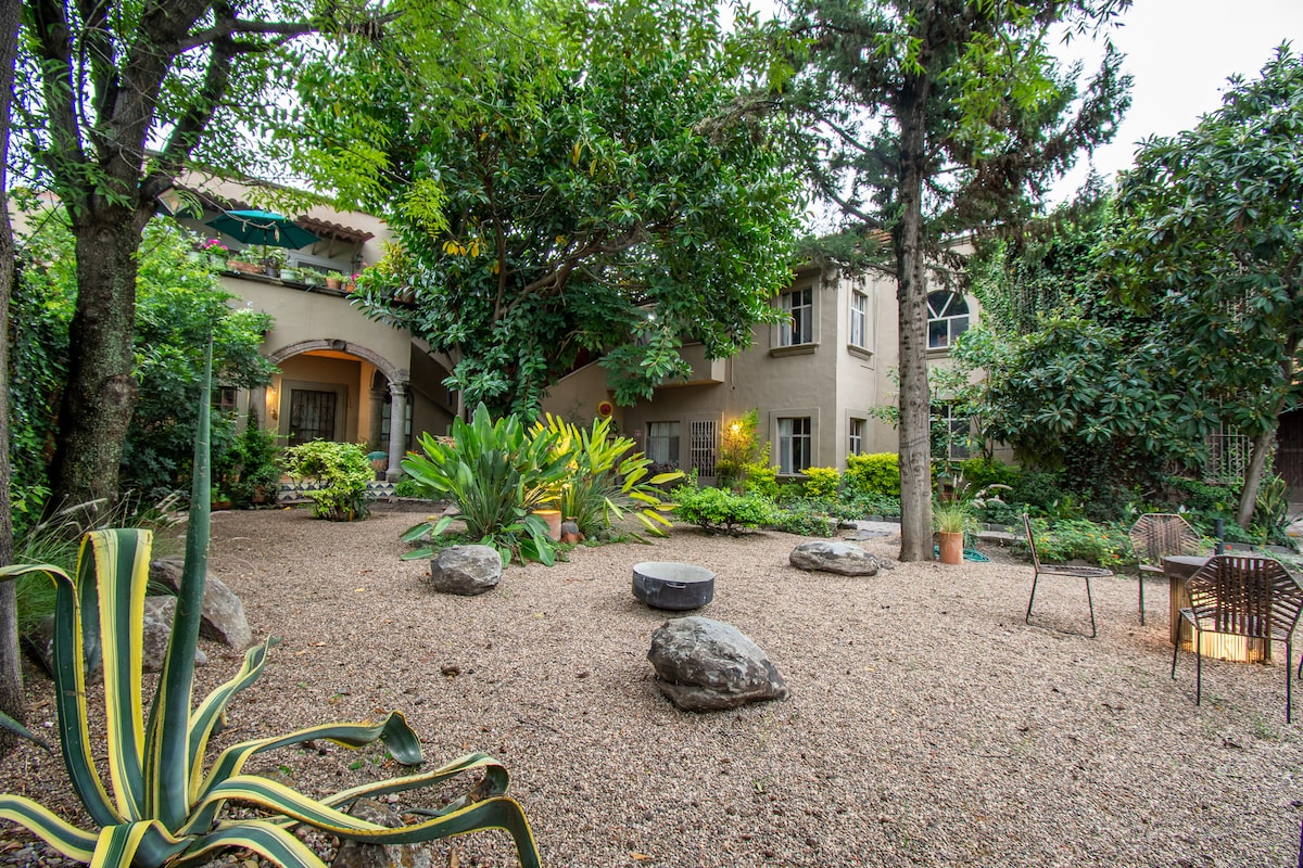 A lush garden area is depicted, filled with greenery including various plants and trees. Smooth stones are scattered throughout the gravel surface, with seating options indicated by chairs positioned around the space. The setting offers a serene atmosphere surrounded by the building's exterior.