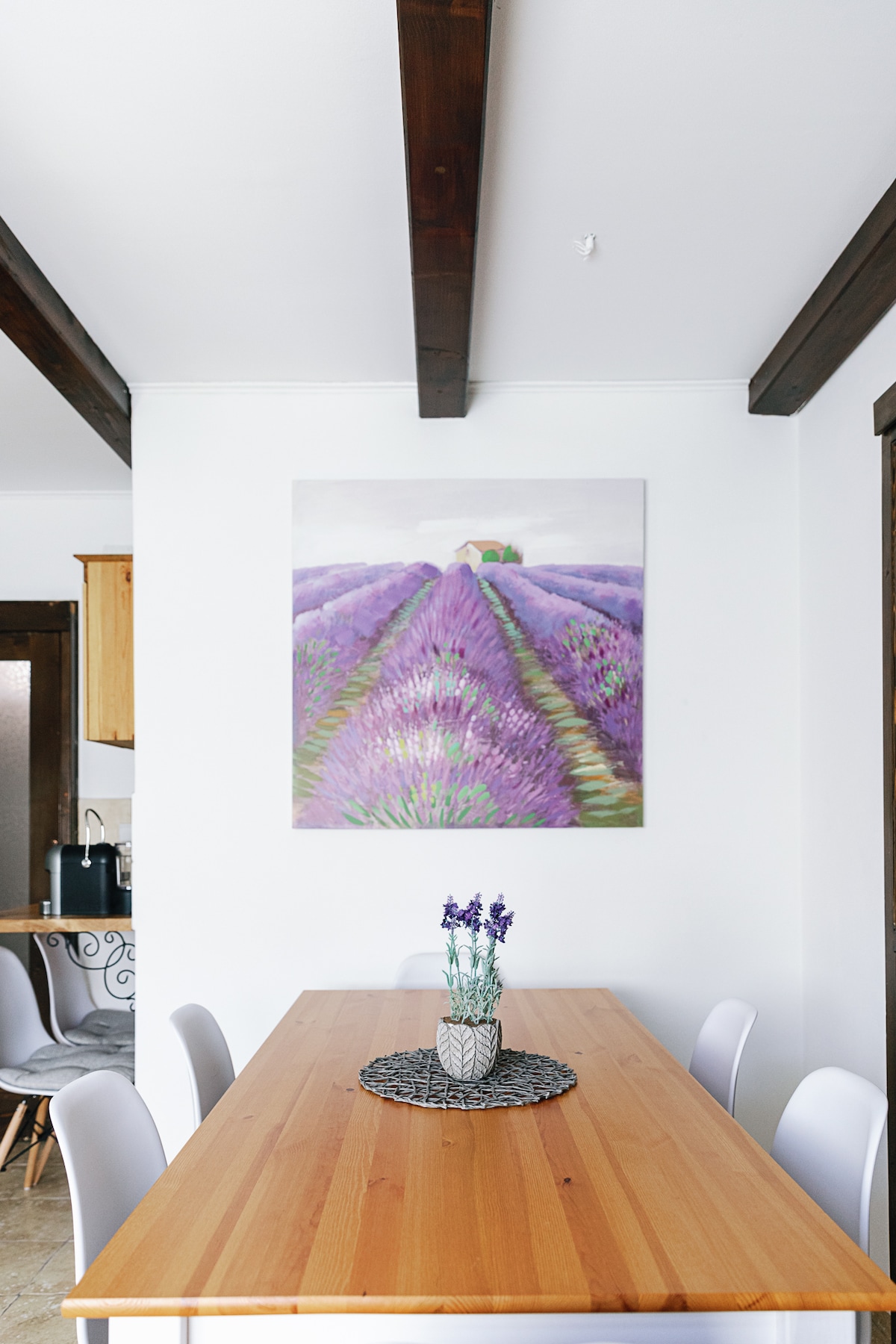 A wooden dining table is centered in the room, surrounded by light-colored chairs. On the table, a vase with lavender flowers is displayed atop a textured placemat. A large artwork depicting lavender fields is mounted on the wall, providing a serene focal point.