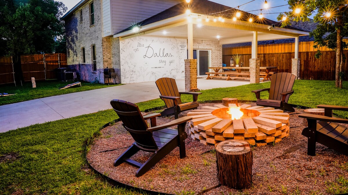 An inviting backyard setting features a fire pit surrounded by rustic wooden seating, creating a cozy atmosphere. String lights hang above, and a spacious covered area offers an oversized picnic table, perfect for gatherings. Lush green grass enhances the outdoor space.