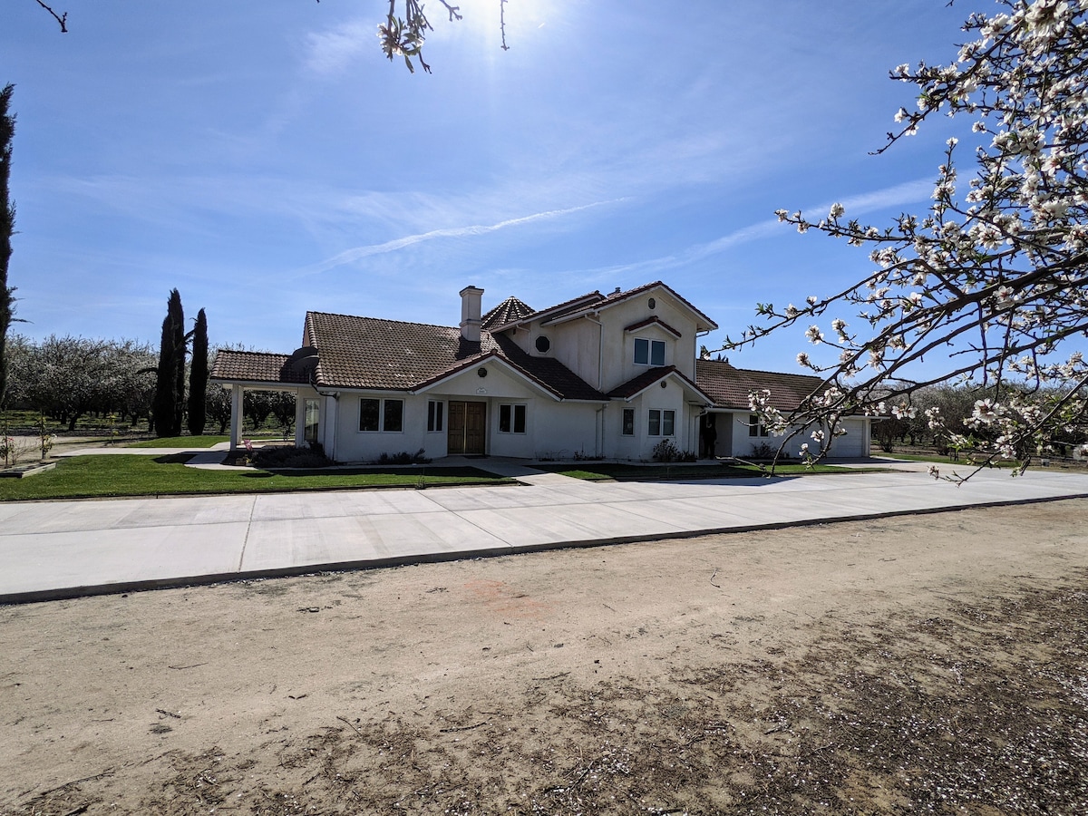 A spacious farmhouse is set against a clear blue sky, surrounded by blooming almond trees. The building features a combination of white and earthy tones, with multiple windows and a large front porch. A broad driveway extends in front of the home, leading to open green areas.