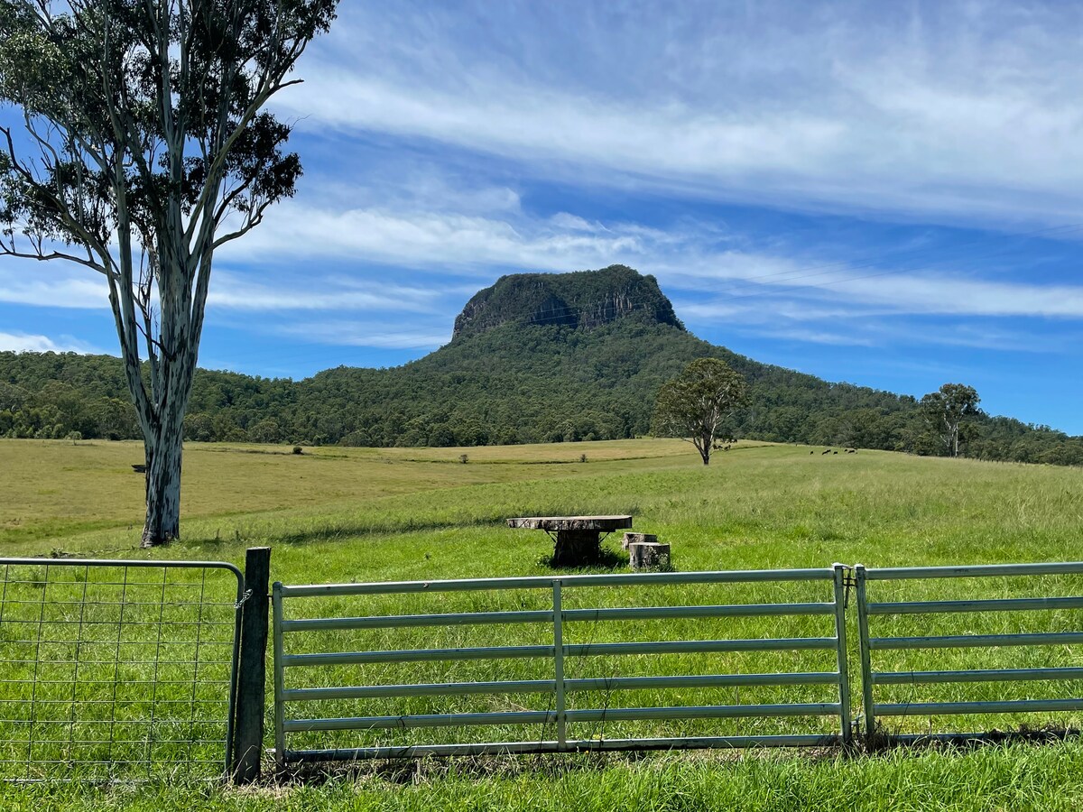An expansive field stretches out, framed by a green fence. In the distance, Mt. Edinburgh Castle rises prominently against a clear blue sky, surrounded by lush greenery. A large flat stone structure can be seen in the foreground, enhancing the tranquil rural landscape.