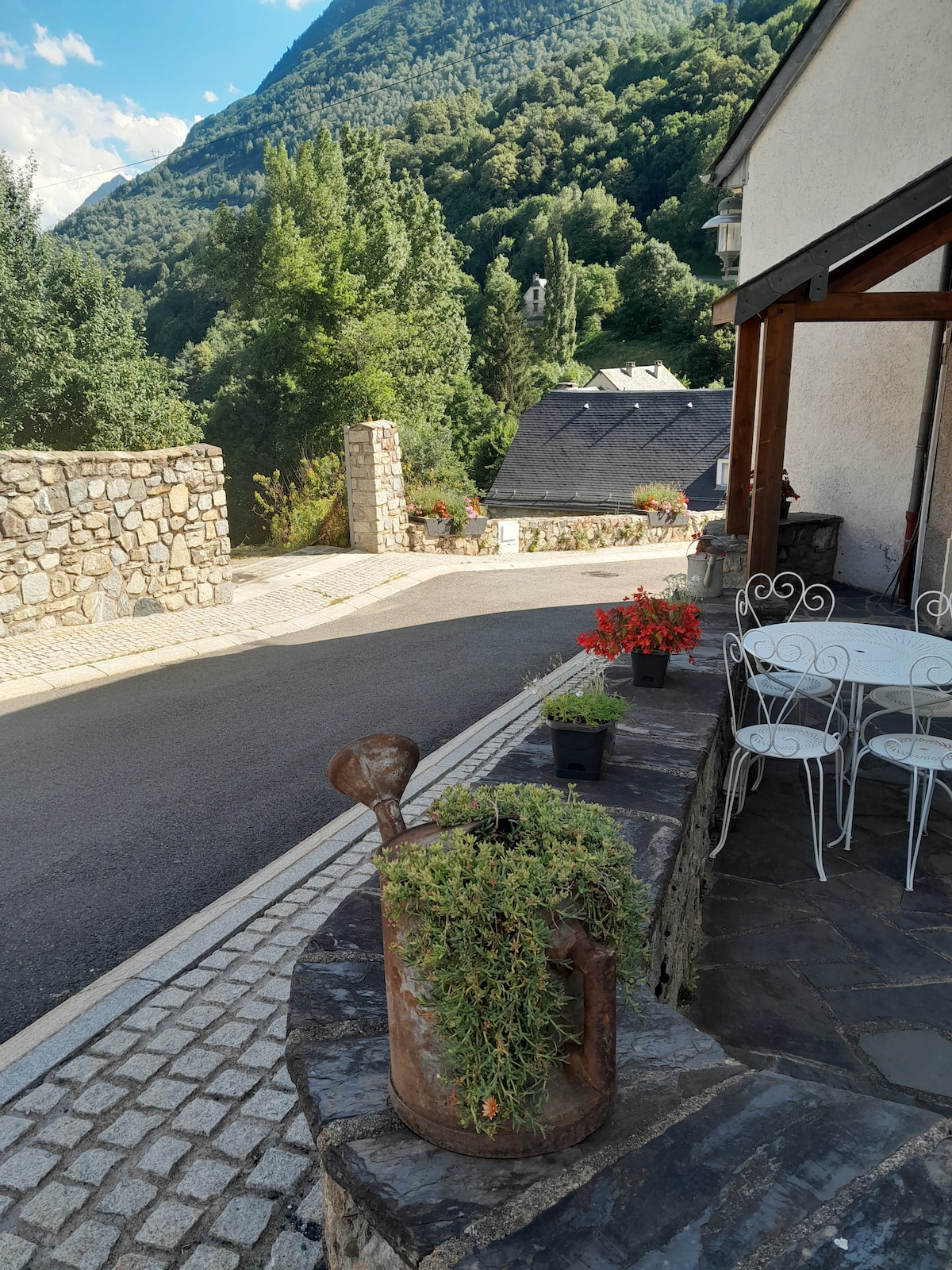 A charming outdoor seating area features a small round table and two metal chairs, positioned alongside a stone wall. An antique watering can filled with greenery adds a natural touch, while the surrounding landscape of hills and trees creates a serene backdrop.