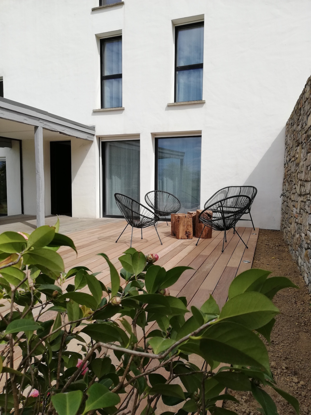 A cozy outdoor seating area is shown, featuring three black wicker chairs arranged around a small wooden table. The space is surrounded by wooden decking and is complemented by greenery in the foreground. Large windows provide natural light and views into the adjoining interior.