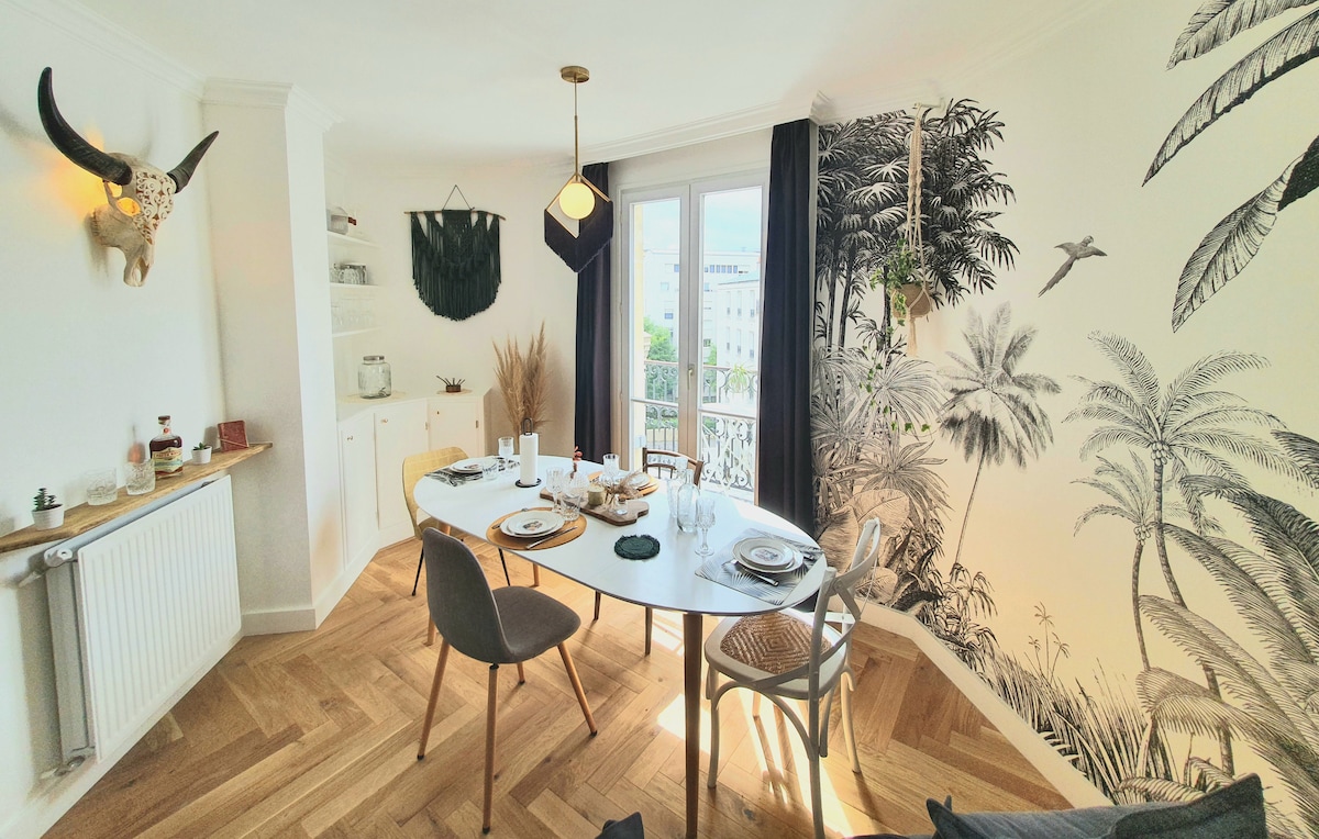 A dining area is presented with a round table set for meals, surrounded by stylish chairs. A wall mural featuring tropical foliage adds a vibrant touch. Large windows allow natural light to fill the space, while decorative elements create an inviting atmosphere.