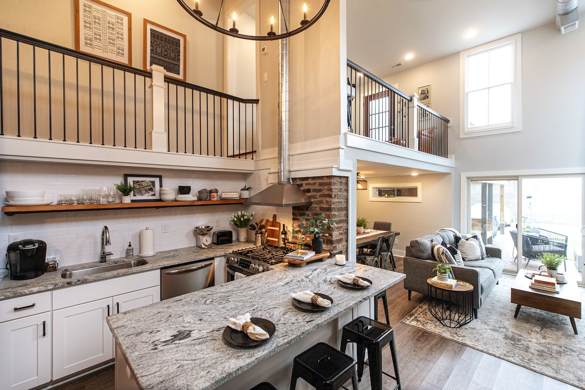 A spacious kitchen with modern appliances features a large island topped with gray stone. Black stools are arranged around the island. A comfortable living area with gray sofas is visible in the background, complemented by large windows allowing ample natural light.