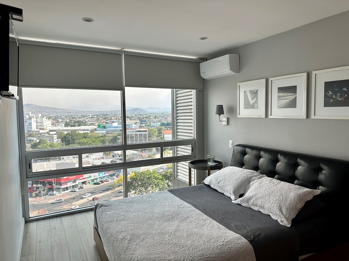 A modern bedroom features a spacious bed with smooth linens, positioned near large windows that offer expansive views of the city. Air conditioning is installed for comfort, while framed artworks adorn the wall, adding a touch of elegance to the space.
