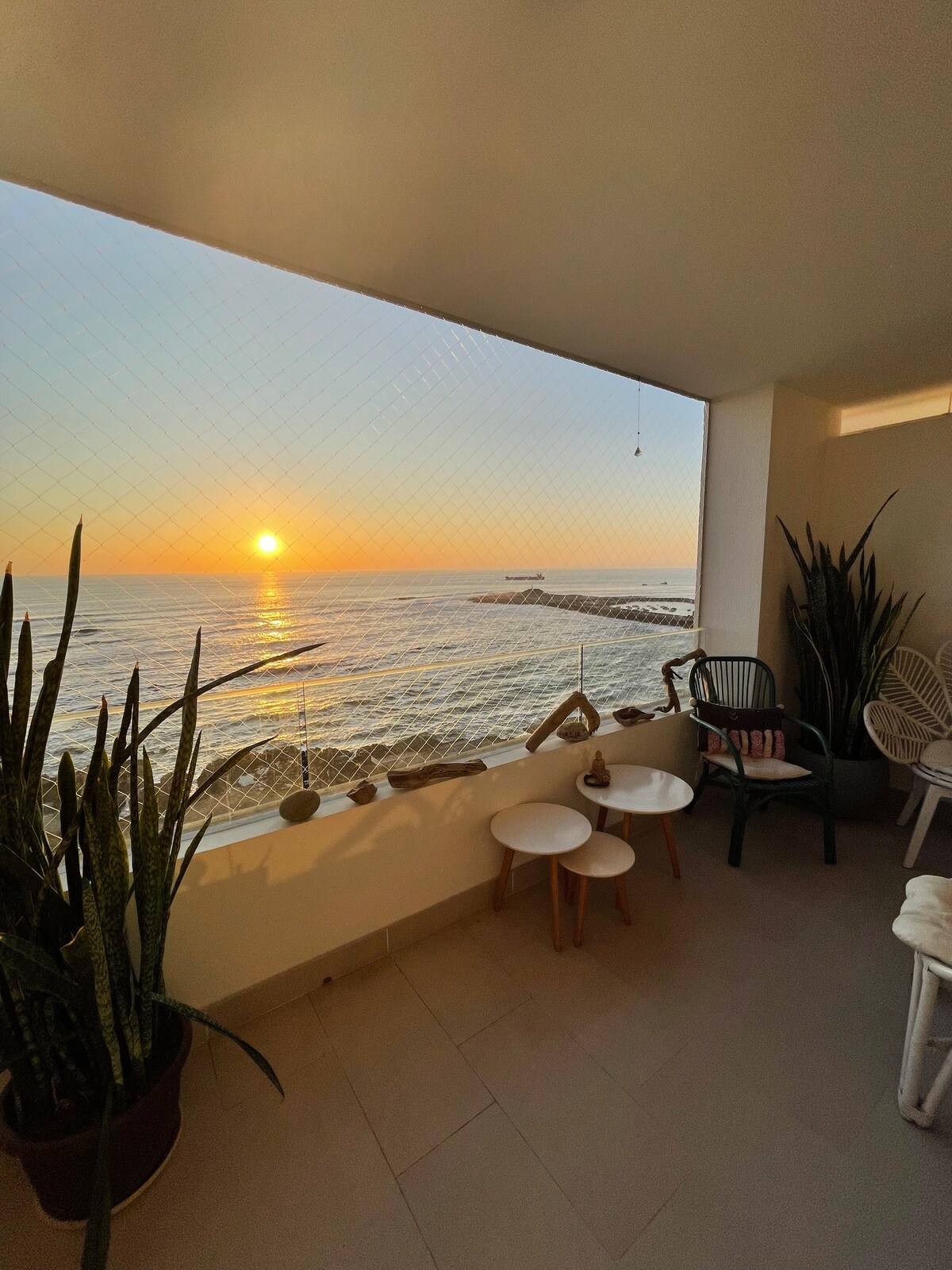 A balcony area is depicted, featuring two small tables beside a selection of chairs. Lush green plants are positioned along the edges, and a sunset is visible over the calm ocean waters, creating a serene backdrop.
