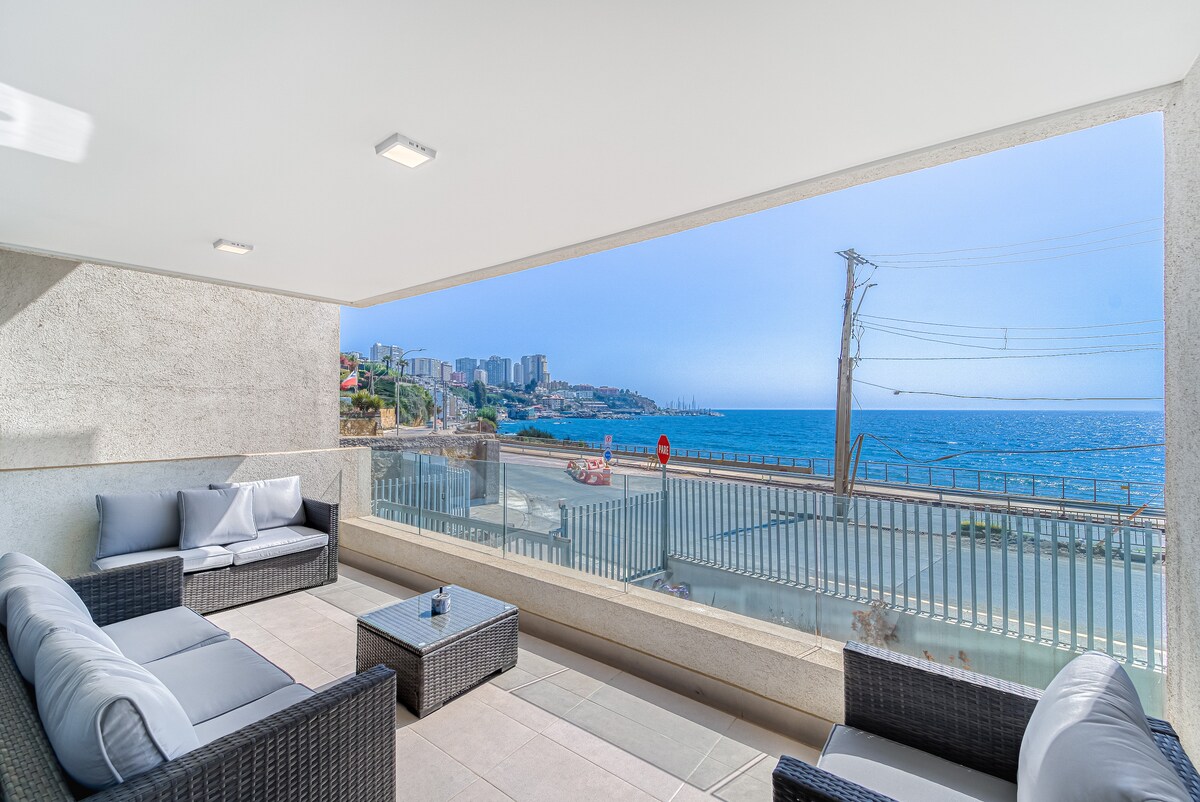 An outdoor terrace features comfortable seating with cushions, offering an unobstructed view of the ocean. The tranquil waters and coastal scenery can be seen through large glass panels, enhancing the connection to the surrounding environment.
