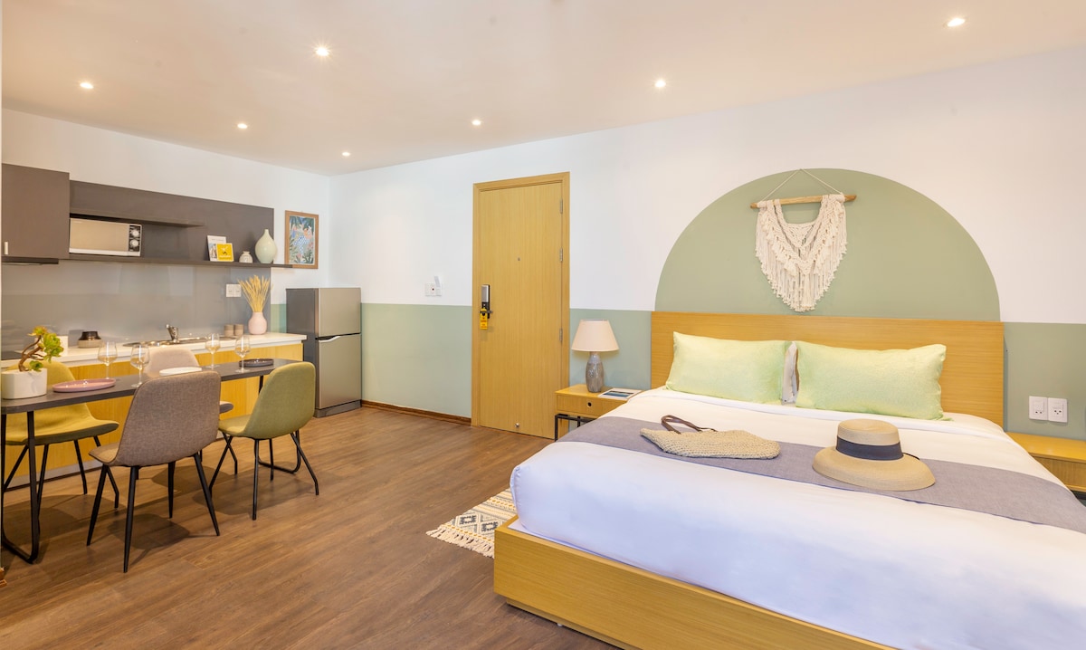 A spacious room features a comfortable king-sized bed with soft linens, complemented by a decorative wall hanging. A dining area with modern chairs and a small table is positioned near a fully equipped kitchenette. Light fixtures provide a warm glow throughout the space.