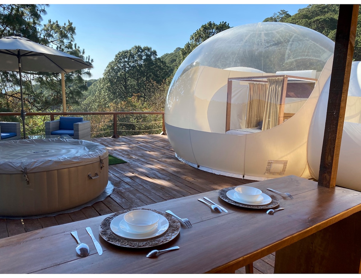 An outdoor dining area features a wooden table set with plates and cutlery, overlooking a wooden deck. A hot tub is situated nearby, while a transparent dome structure is visible in the background, set amidst lush greenery and trees.