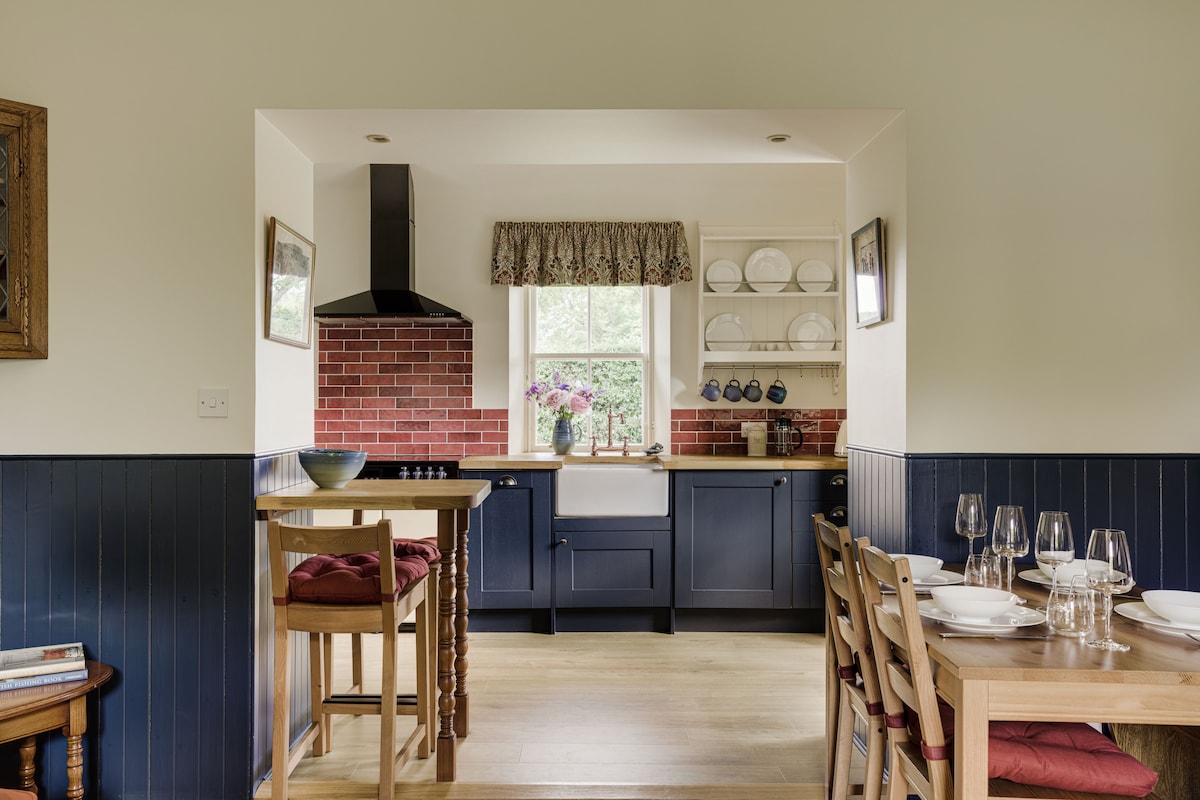 An open-plan kitchen and dining area features dark blue cabinetry and red-brick accents. A wooden dining table with placed settings sits adjacent to bar stools. Shelves with dishware and a window adorned with floral curtains allow light to fill the space.